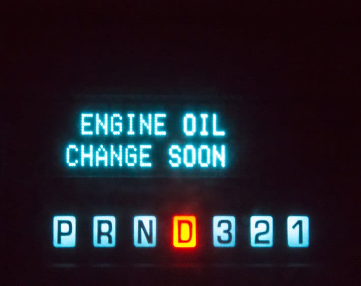 Lights on a car dashboard at night that says "Engine oil change soon". The words are in blue. The symbols for what gear the automobile is in is also lit. The letter D is orange, showing that the vehicle is in drive.